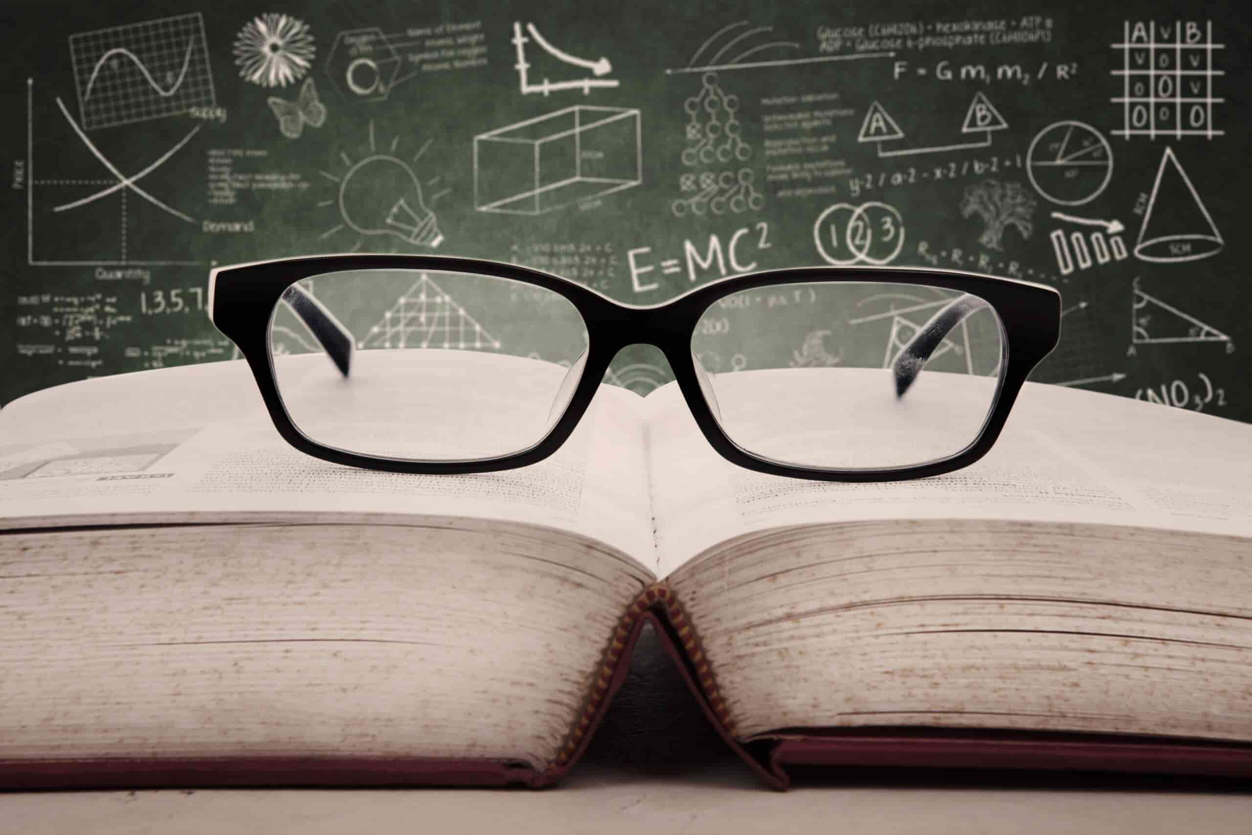 A pair of black glasses sits atop an open book. In the background, a chalkboard displays various mathematical and scientific formulas and diagrams, including graphs, a cube, and the equation E=MC².