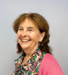 A woman with shoulder-length brown hair smiles at the camera. She is wearing a colourful floral blouse and a pink cardigan. The background is plain and light grey.