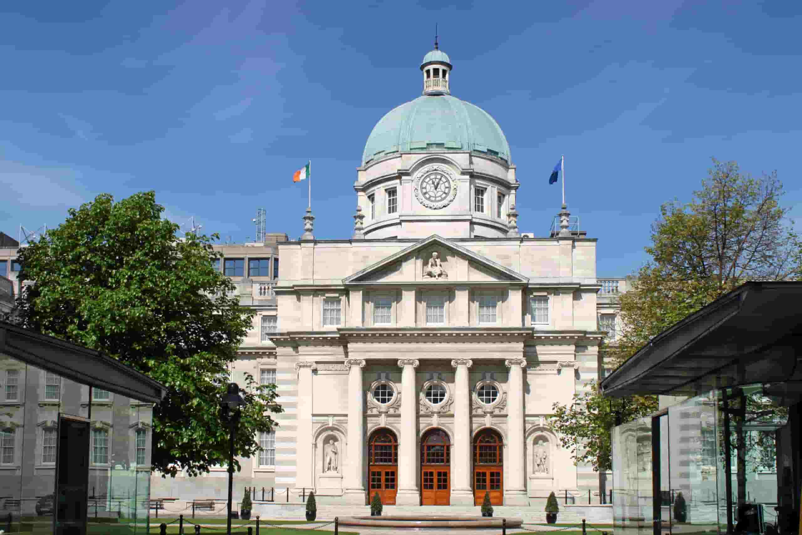 Government Buildings Dublin, a building with a prominent dome and clock stands under a clear blue sky. Two flags are flown, and lush green trees frame the entrance with three arched wooden doors.