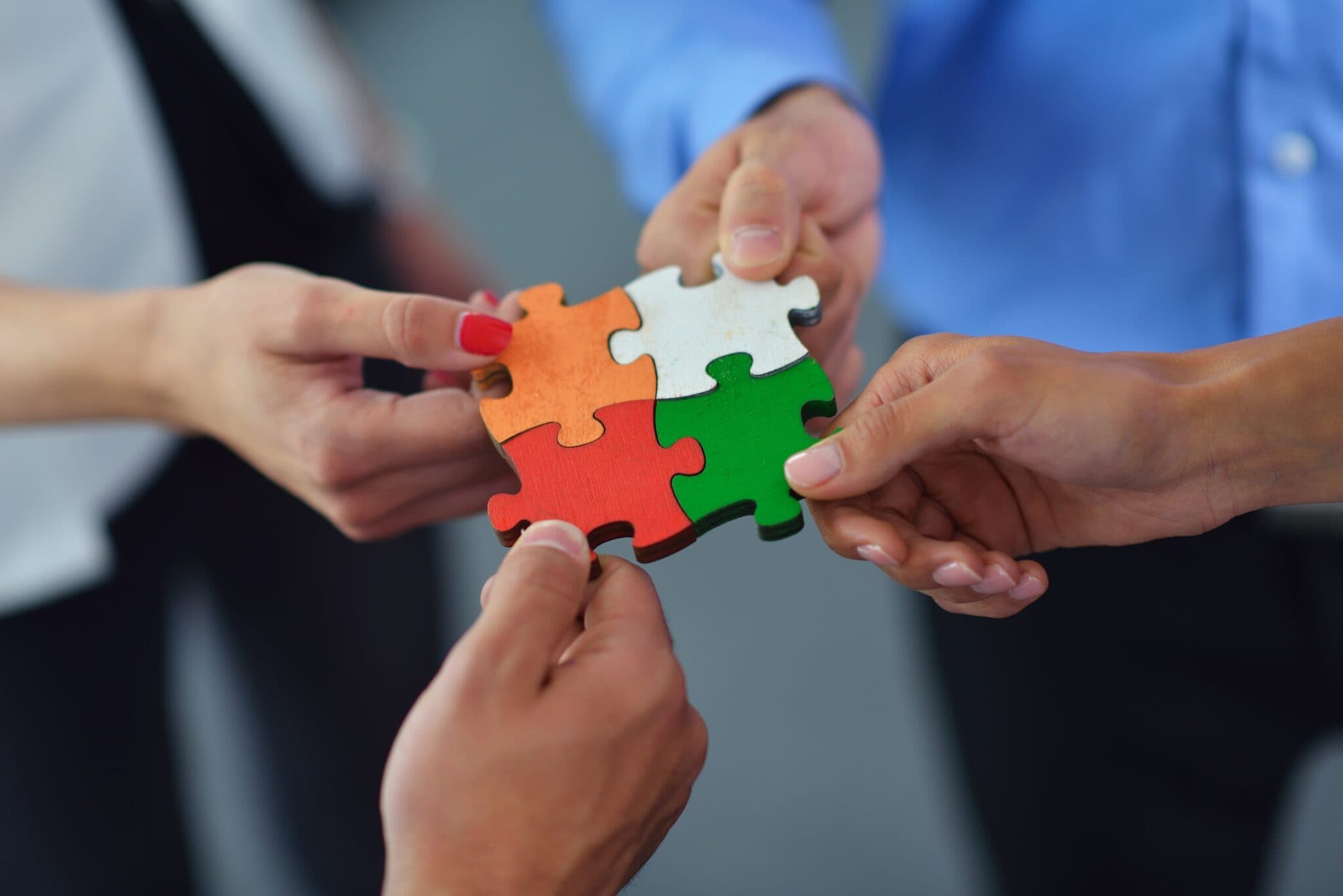 Four hands holding colourful jigsaw puzzle pieces fitting together. Each piece is a different colour: orange, white, green, and red, symbolising teamwork and collaboration. The hands belong to people wearing business attire.