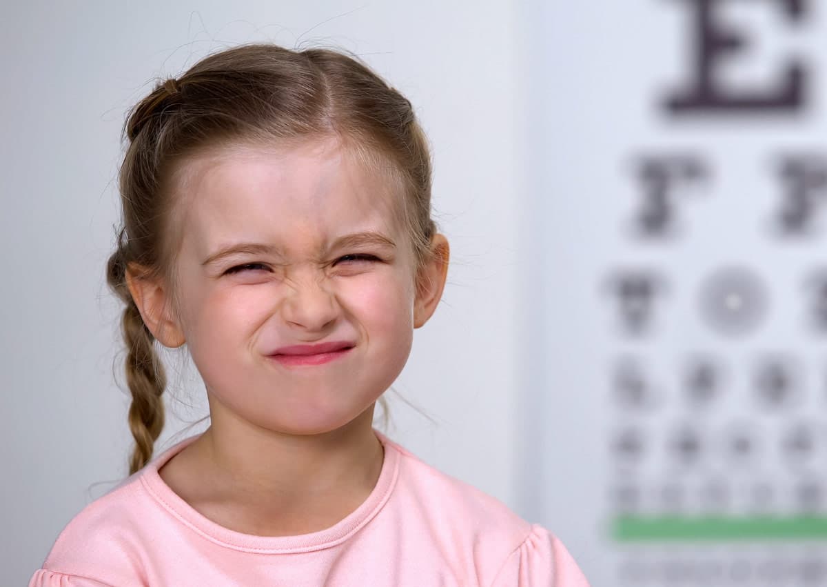 A young girl squints her eyes while standing in front of an eye chart. She has braided hair and wears a light pink shirt. The eye chart is slightly blurred in the background.