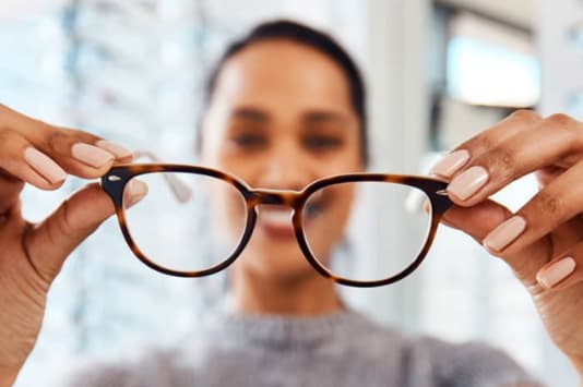 A person holds a pair of tortoiseshell glasses in front of them, focusing on the lenses. The background is softly blurred, highlighting the eyewear with a light and neutral setting. The person’s fingernails are painted light pink.