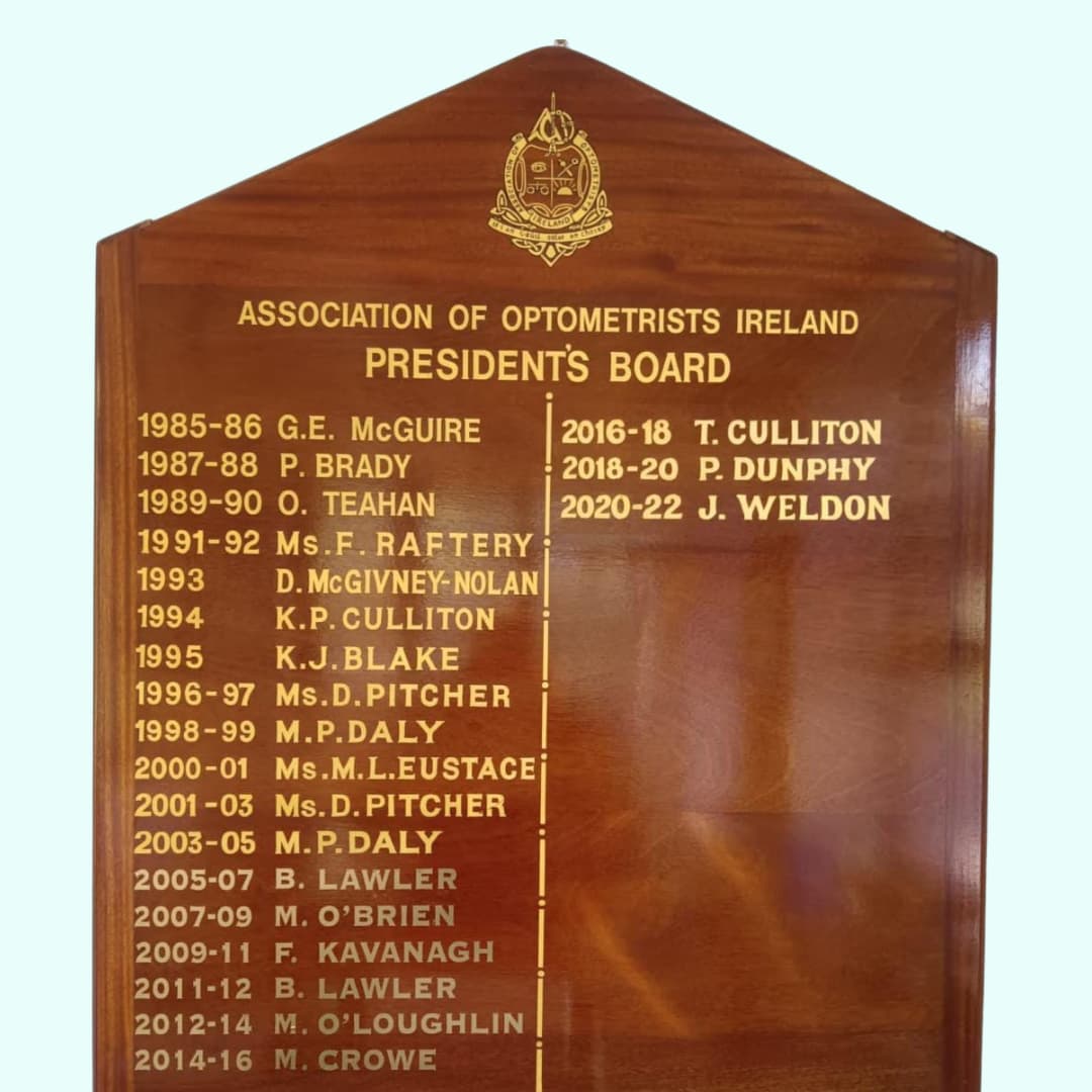 A wooden plaque titled Association of Optometrists Ireland Presidents Board lists names and terms of presidents from 1985 to 2022. The board has a polished finish and an engraved emblem at the top.