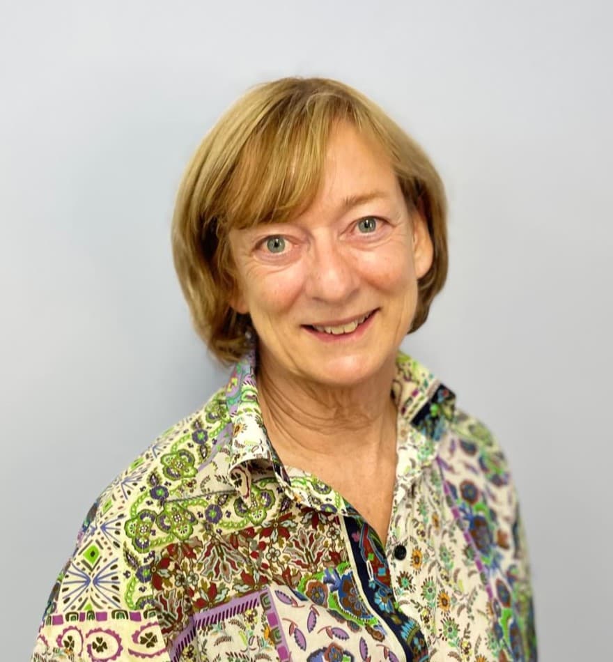 A smiling woman with short, light brown hair wearing a colorful, patterned blouse stands against a plain background.