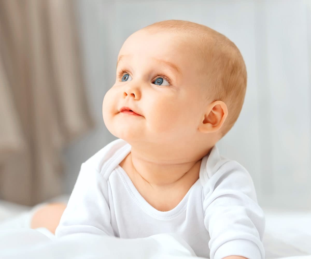 A smiling baby in a white outfit is lying on their stomach, looking upwards. The background is softly blurred, giving a cozy and serene atmosphere.