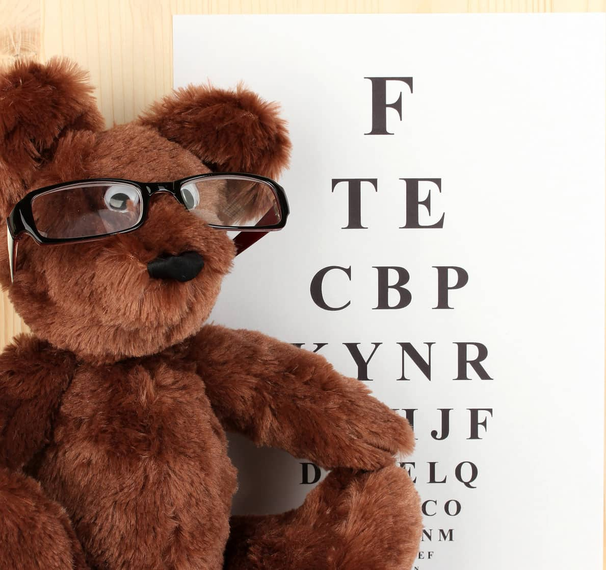 A brown teddy bear wearing glasses sits in front of an eye chart, which displays letters of varying sizes. The bear appears to be leaning against a light wooden surface.