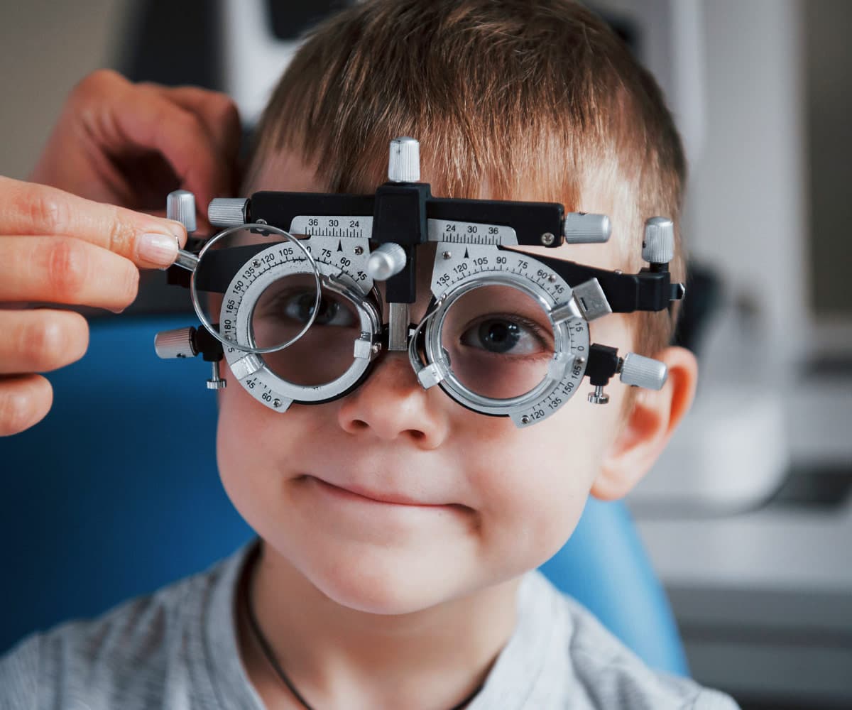 A young child sits in a vision testing chair wearing trial frames for an eye exam. An adults hands adjust the frames to fit properly. The child looks ahead with a slight smile.