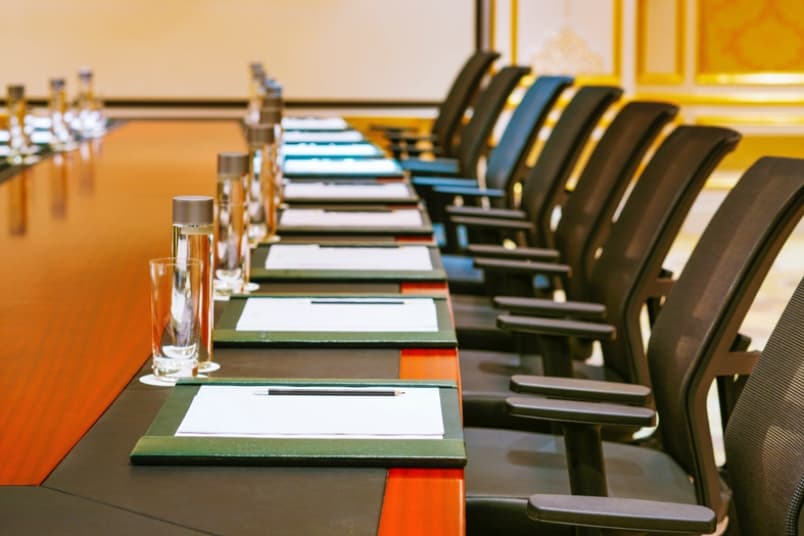 A long conference table is lined with black office chairs. Each place is set with a notepad, pen, and a water bottle. The room has elegant decor with gold accents.