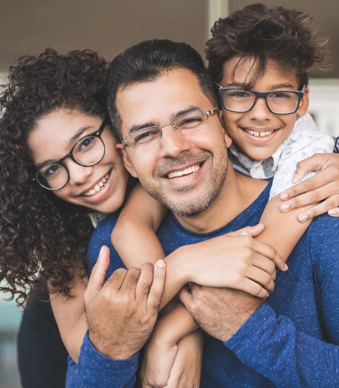 A smiling man wearing glasses is hugged by two children, a girl on his right and a boy on his left. They are all wearing glasses and have curly hair. The children are smiling and leaning affectionately on his shoulders.