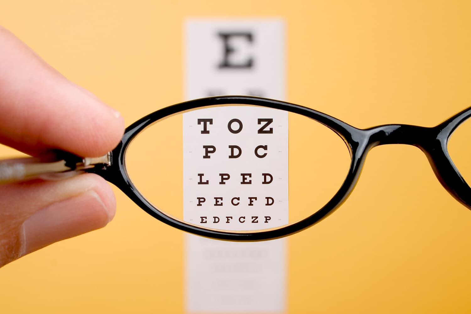 A hand holds a pair of glasses in front of an eye chart with letters of varying sizes. The glasses lens brings the chart into sharp focus, highlighting the top row of letters. The background is a solid yellow color.