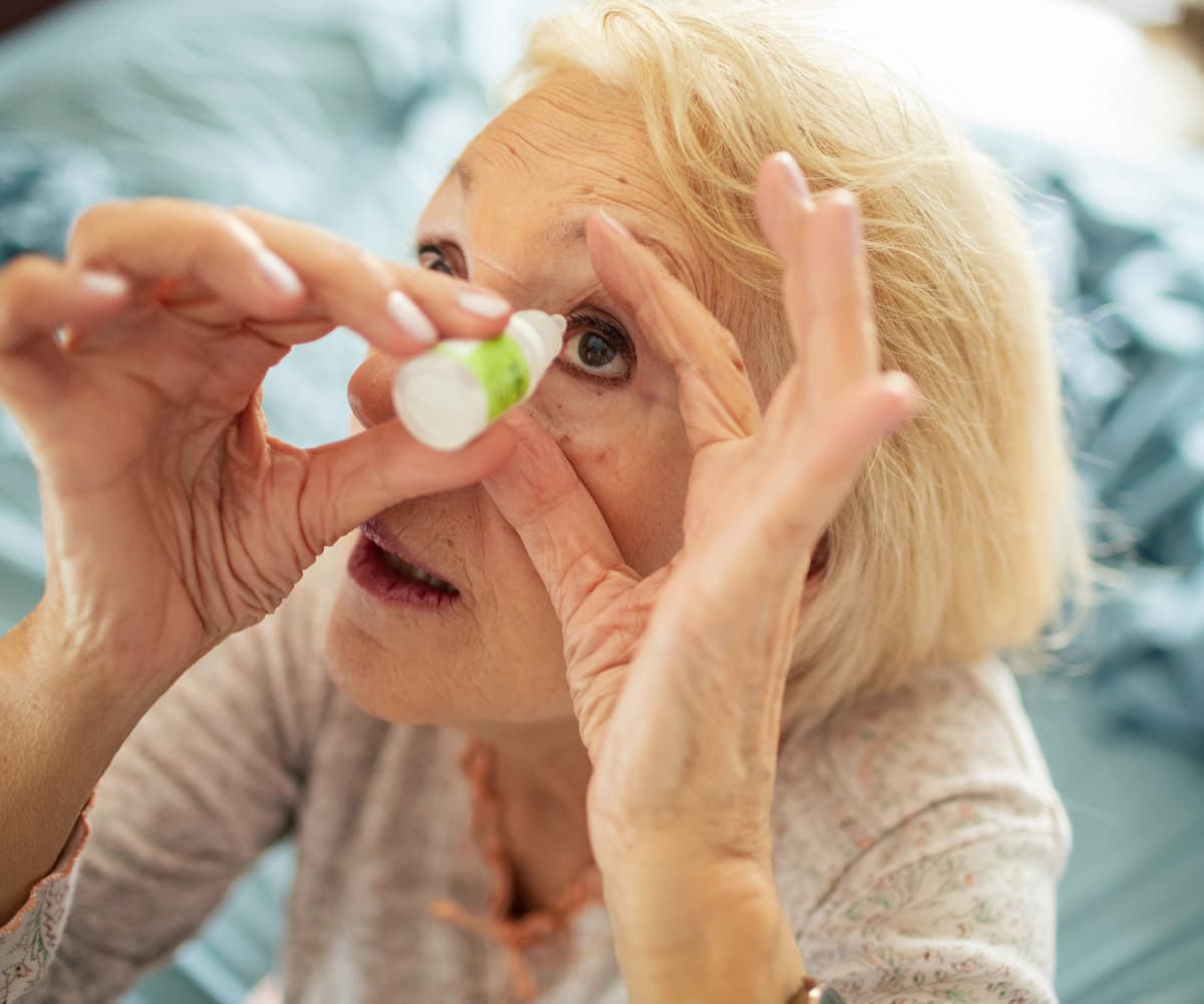 An elderly person with short blonde hair is tilting their head back, applying eye drops into their right eye. The background is blurred, and they appear to be in a home setting, wearing a light-coloured shirt.