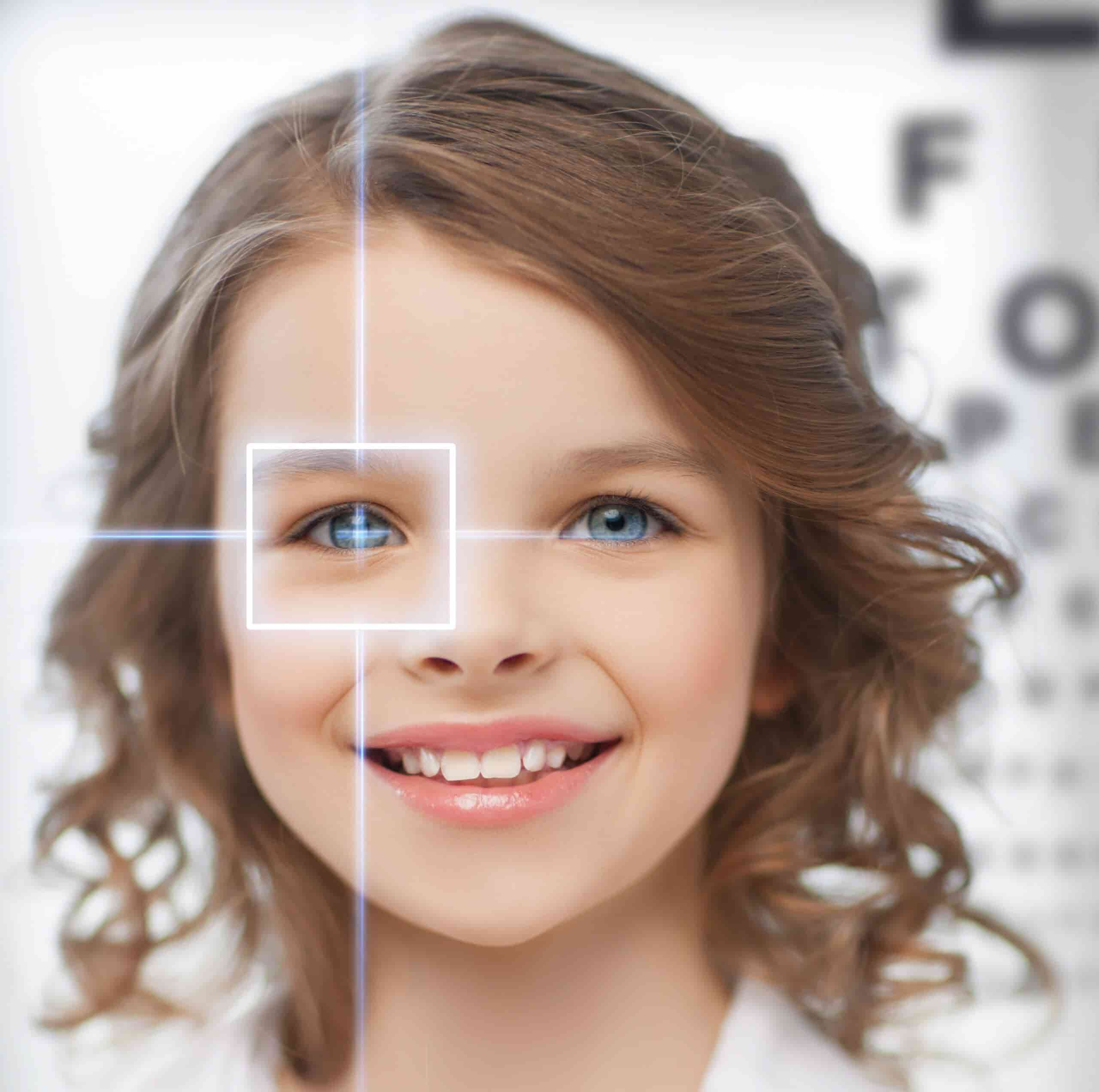 A young girl with curly brown hair and blue eyes smiles. There's a white geometric square overlay on her left eye, with an eye chart  in the background.