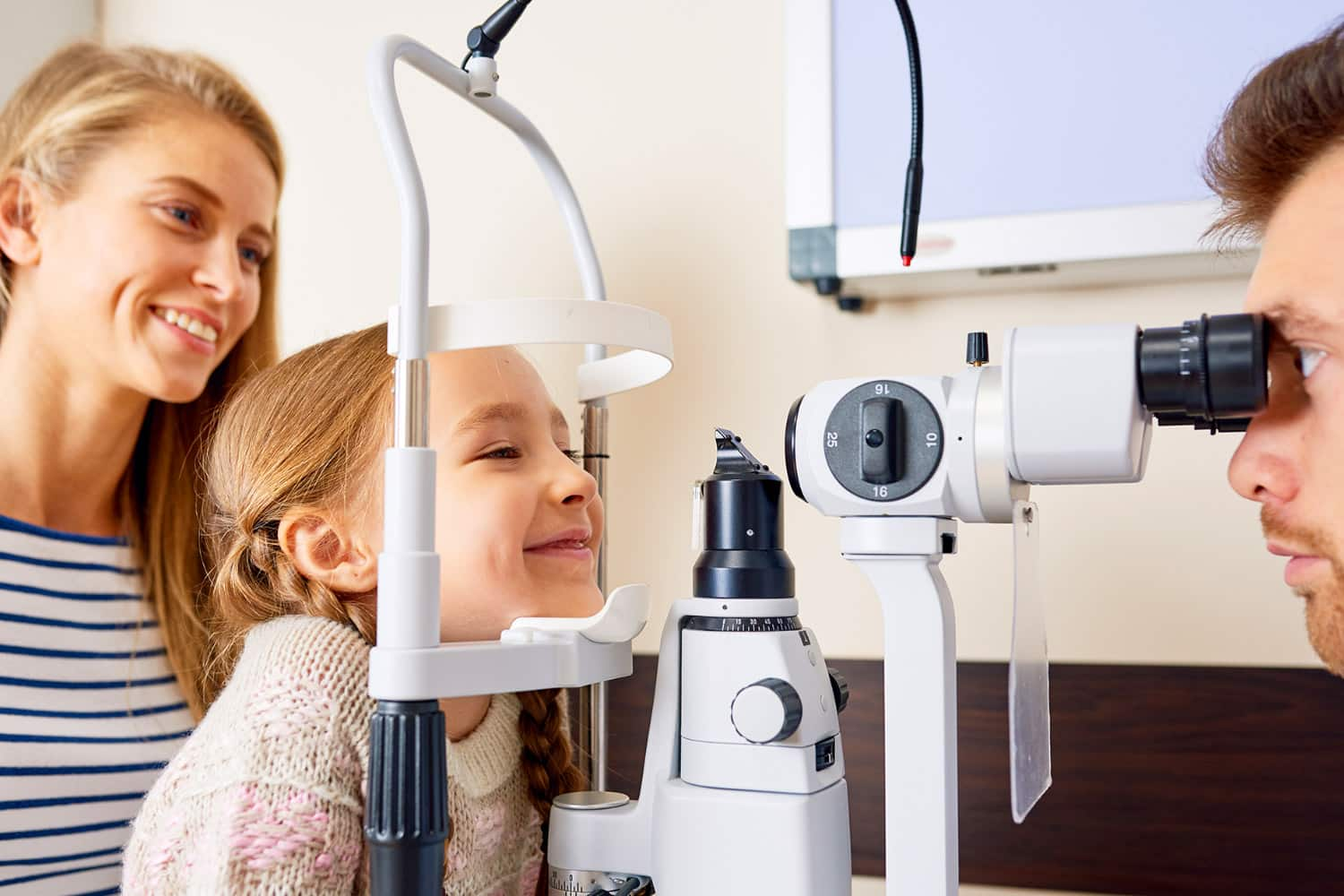 A young girl sits at an eye examination machine, smiling as she looks through it. A woman, possibly her mother, stands beside her, smiling. An optometrist adjusts the machine, focused on the girls eyes.