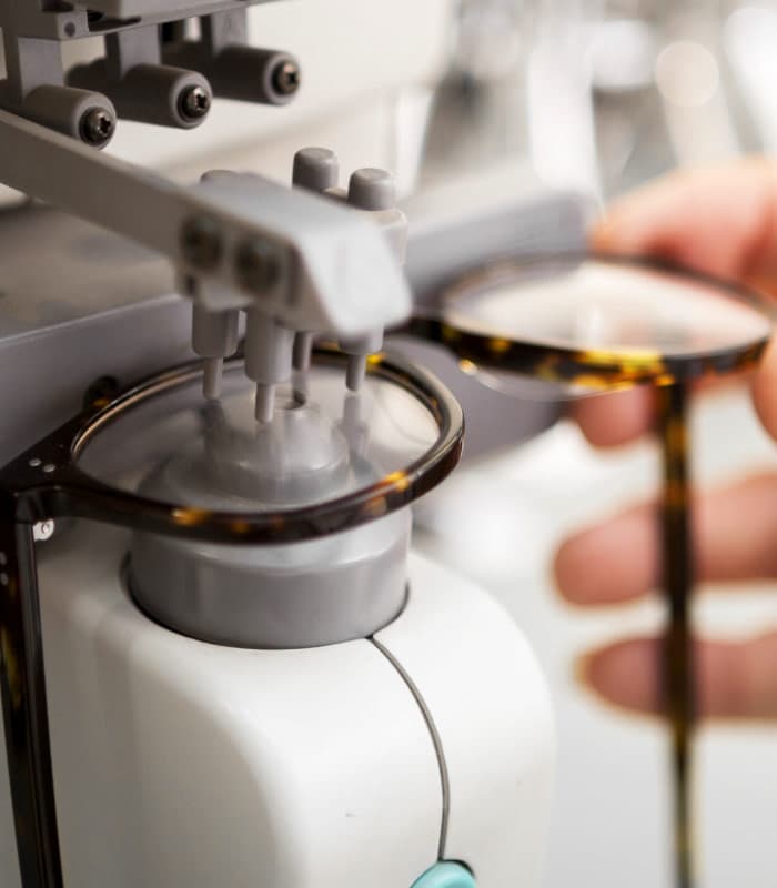 Close-up of a pair of eyeglasses being held by a hand under a lens cutting or polishing machine, showcasing the process of preparing glasses.