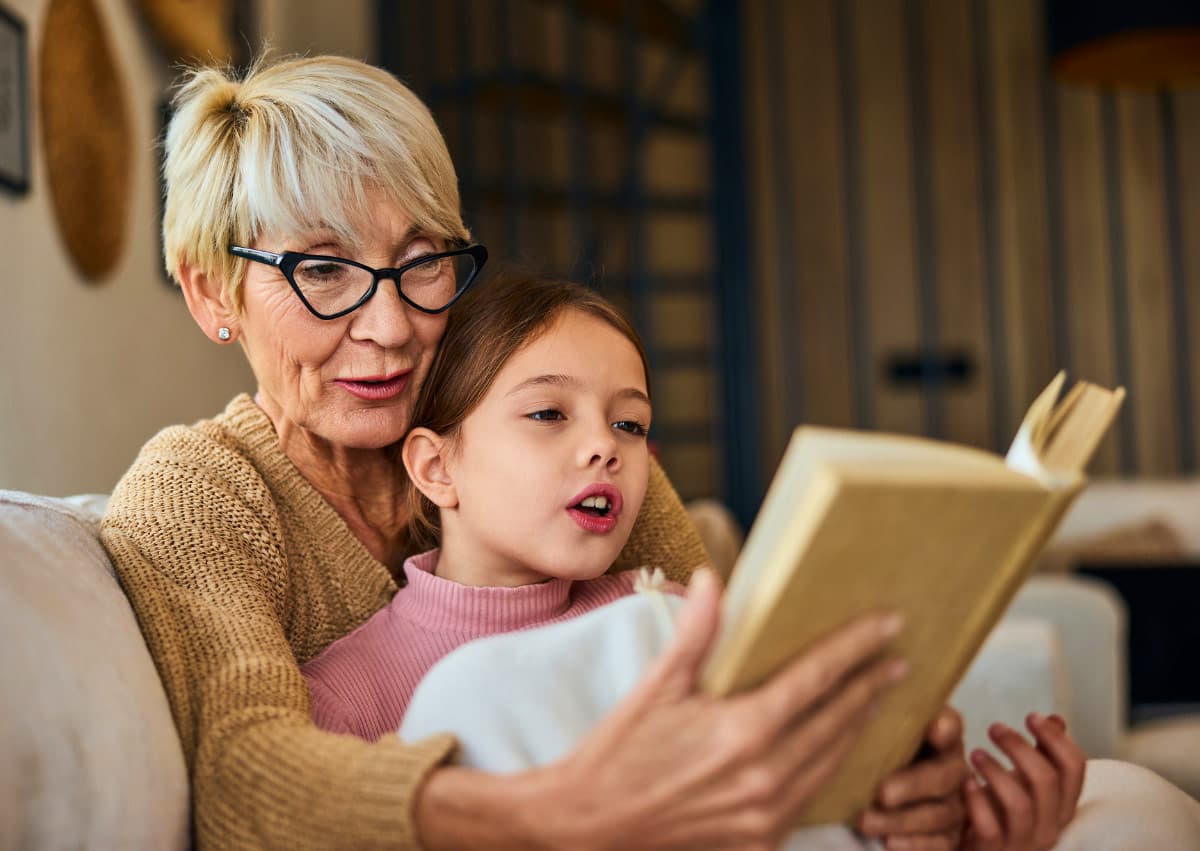 Elderly woman with glasses and short hair is reading a book with a young girl in a pink sweater. They are sitting on a sofa, wrapped in a blanket, with warm lighting creating a cozy atmosphere.