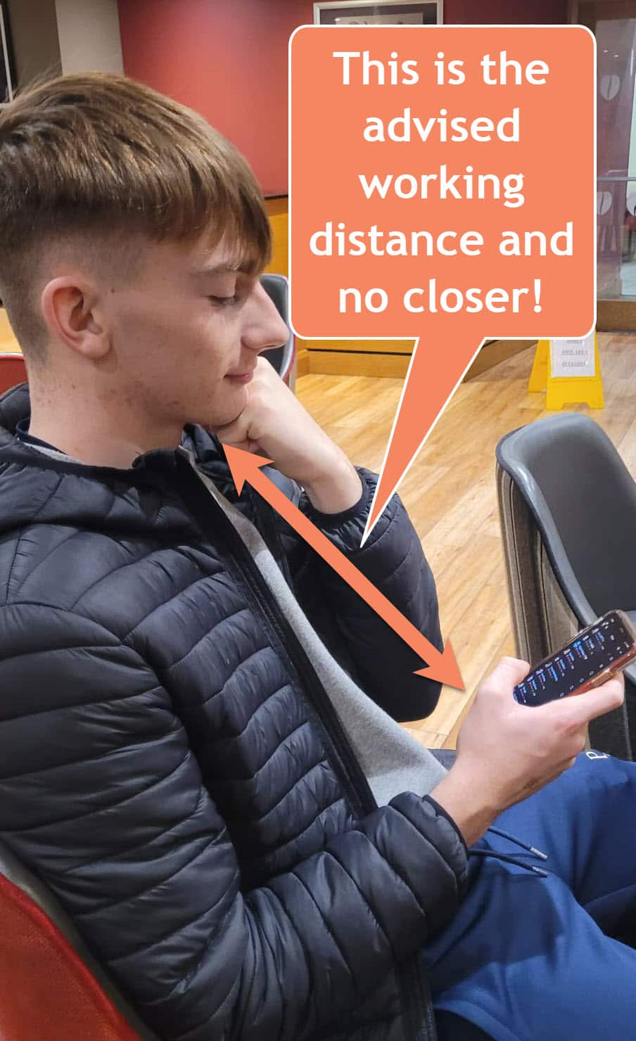 A young person in a black jacket looks at their phone, which is held at eye level. An arrow points to the phone with text that reads, This is the advised working distance and no closer! The background shows a red and beige interior.