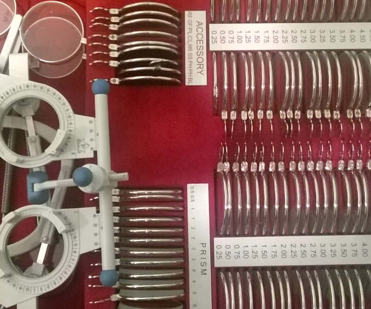 An assortment of optometry trial lenses and prisms are organised in a case. The lenses are lined up in pairs of different strengths. On the left, a trial frame is visible, used for fitting lenses during eye exams.