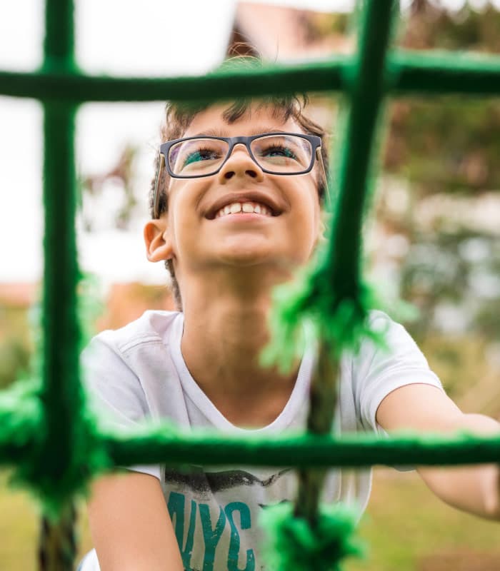 A child wearing glasses and a white t-shirt smiles while playing on a green rope climbing structure outdoors. The focus is on the joyful expression, with the background softly blurred.