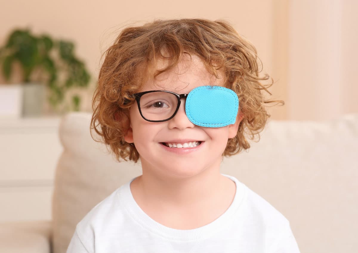 Young child with curly hair and glasses, wearing a blue eye patch over one eye, smiles while sitting indoors. A plant is in the background.