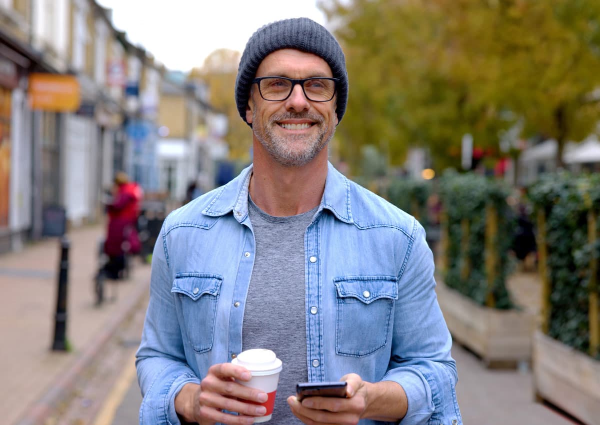 A man wearing a beanie, glasses, and a denim jacket smiles while holding a smartphone and a coffee cup. He is walking along a tree-lined street with shops in the background on an autumn day.