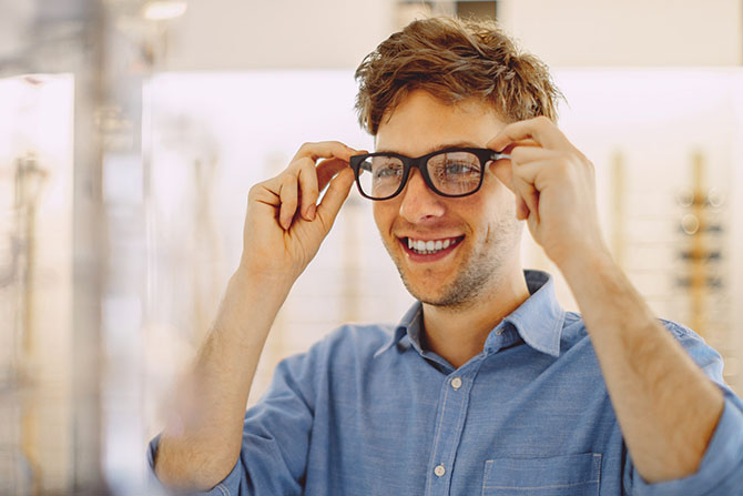 A smiling man in a blue shirt tries on a pair of black eyeglasses in a bright store.