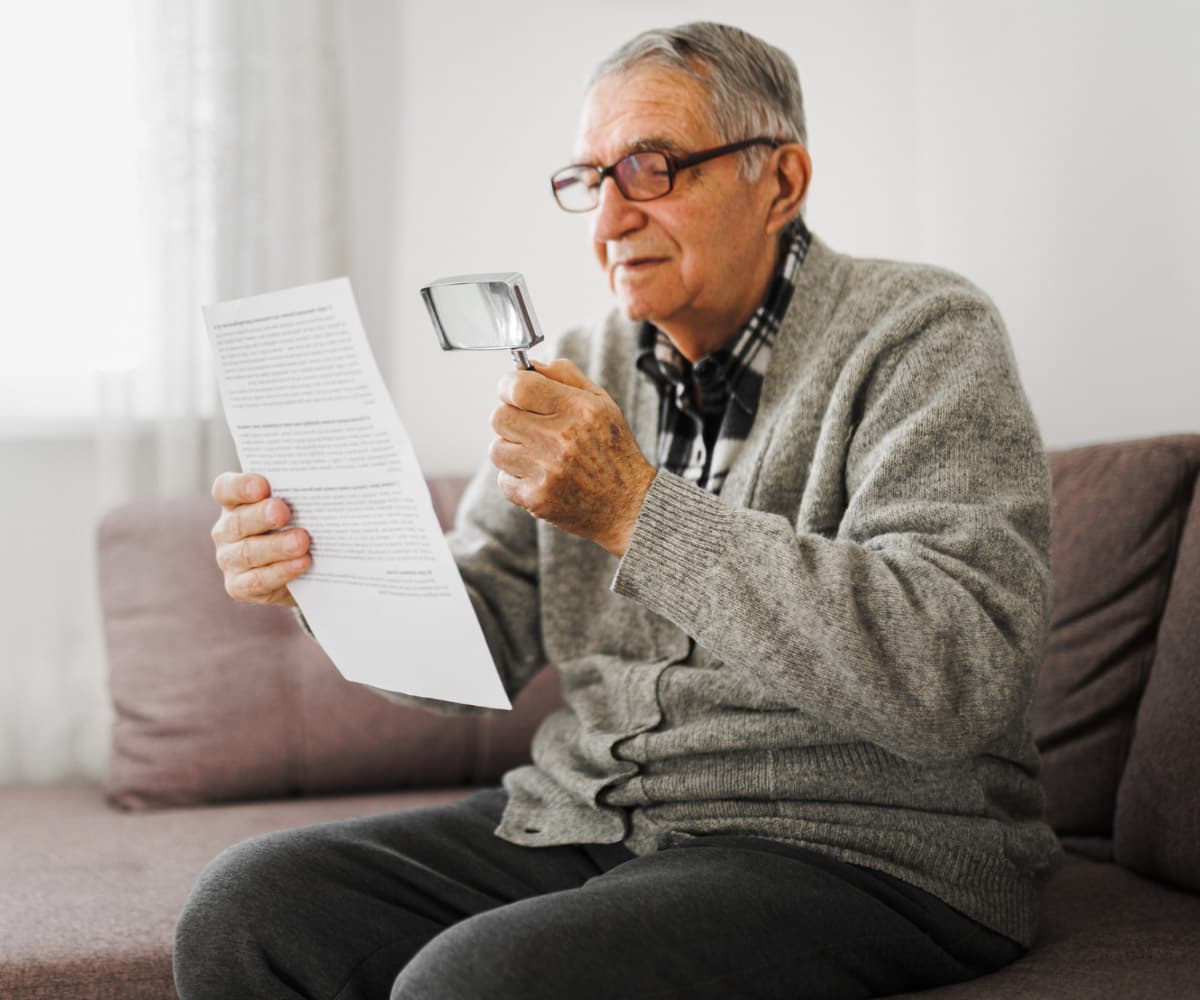 An elderly man with glasses sits on a couch, holding a magnifying glass in one hand while reading a document in the other, symbolising age-related macular degeneration (AMD). He wears a grEy sweater over a plaid shirt, focusing intently on the paper.