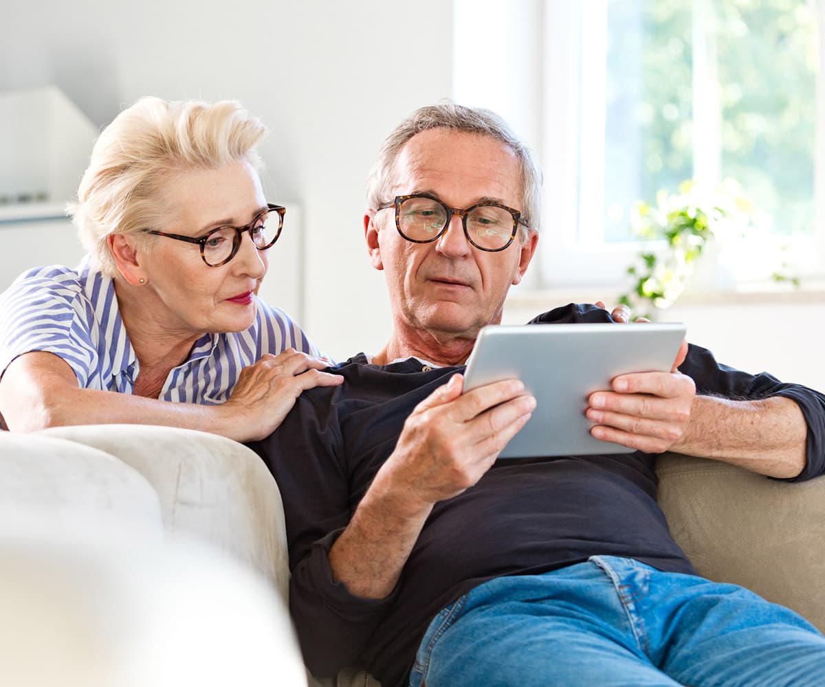 An older couple sits on a sofa, looking at a tablet. The woman, wearing glasses and a striped shirt, leans on the man's shoulder. The man, also wearing glasses, holds the tablet, focused on its screen. Sunlight filters through a window behind them.