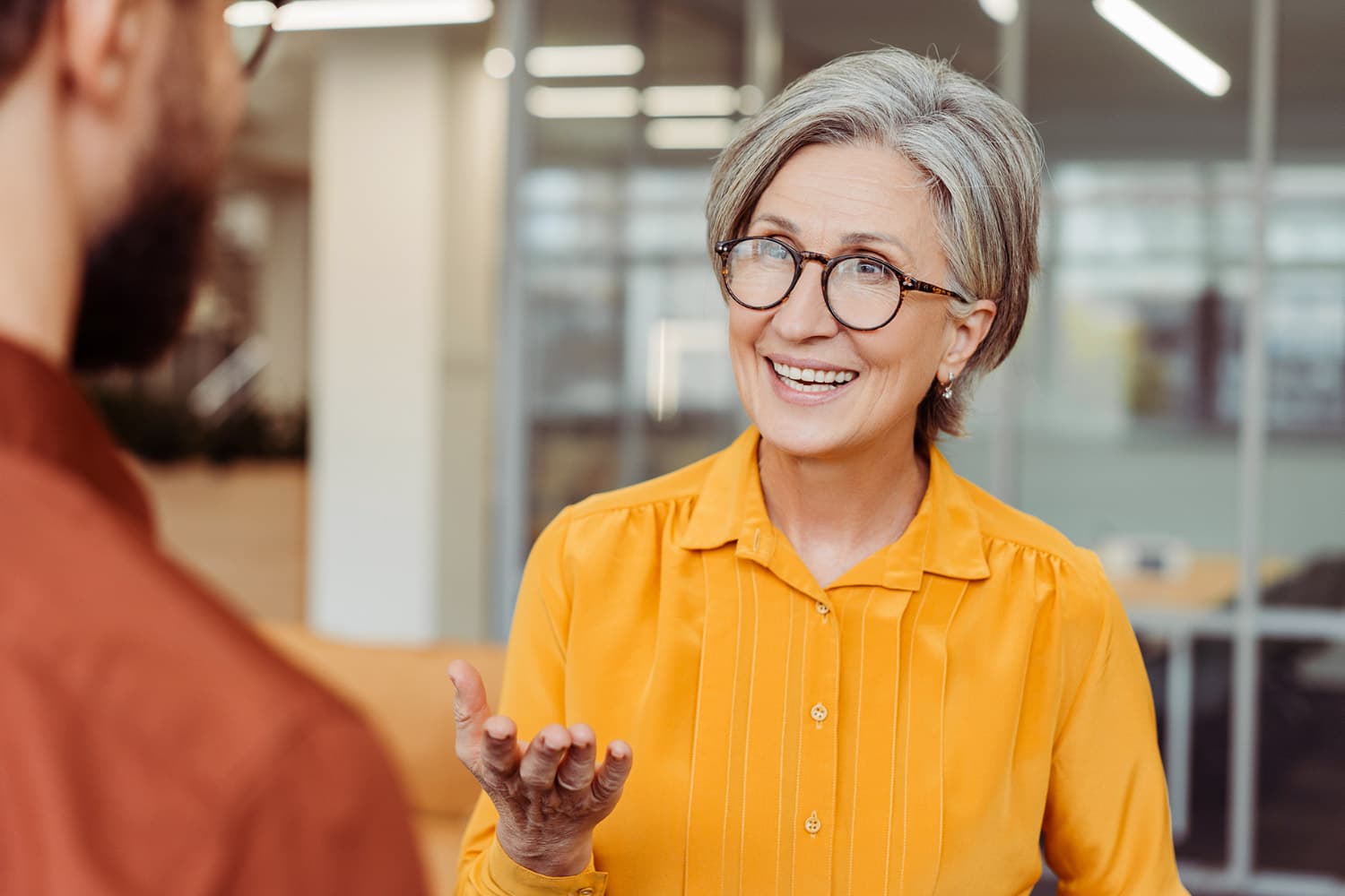 An older woman with grey hair and glasses, wearing a yellow shirt, is smiling and talking to a man with a beard in an office setting. She has her hand raised slightly, and the background shows office furniture and windows.