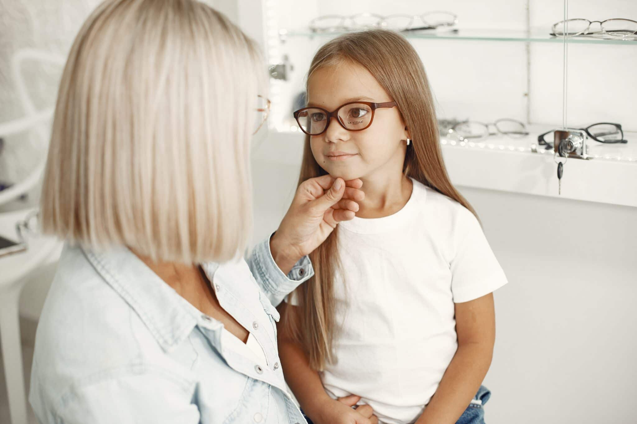 A woman with blonde hair and glasses is adjusting the glasses on a young girl with long brown hair. They are in an eyewear store with shelves of glasses in the background. The woman is wearing a denim shirt, and the girl is in a white t-shirt.