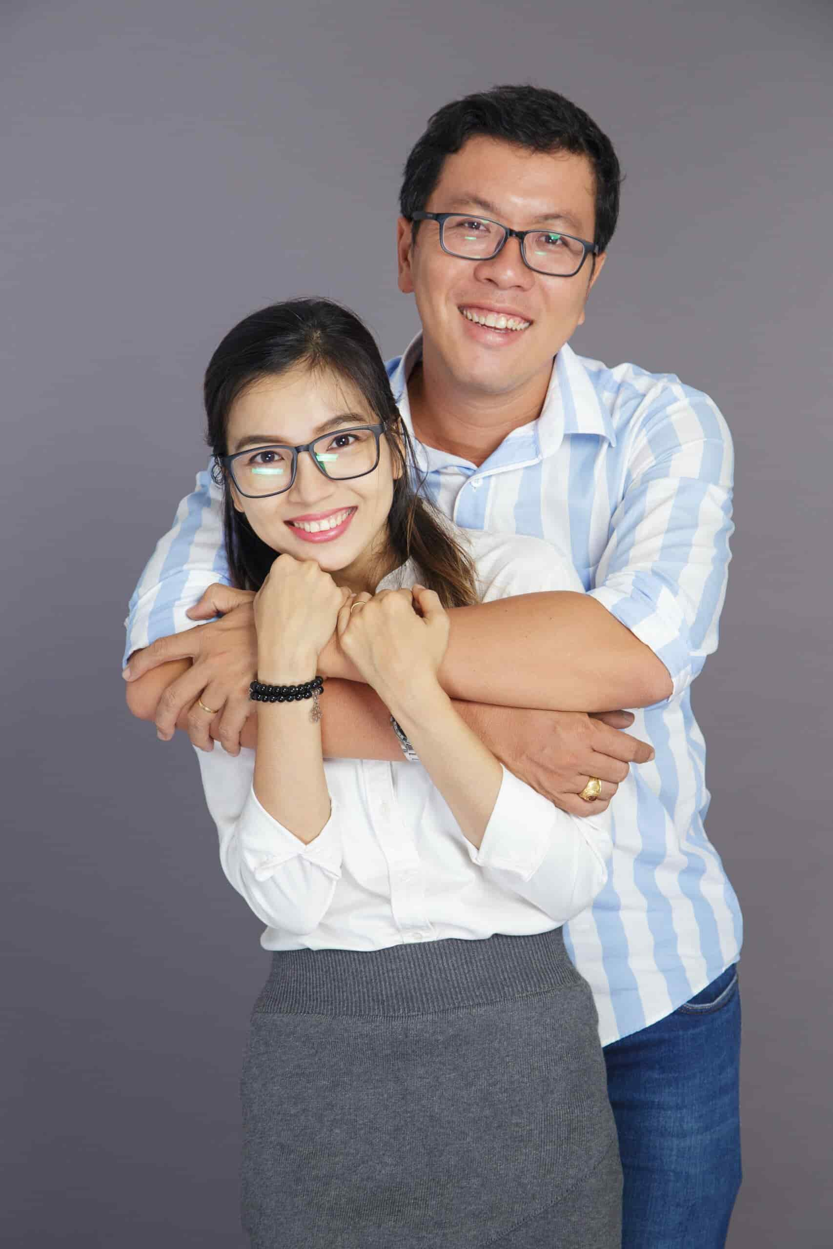 A man and woman wearing glasses stand smiling against a grey background. The man is wearing a striped shirt and embraces the woman from behind. The woman is wearing a white blouse and grey skirt. Both appear happy.