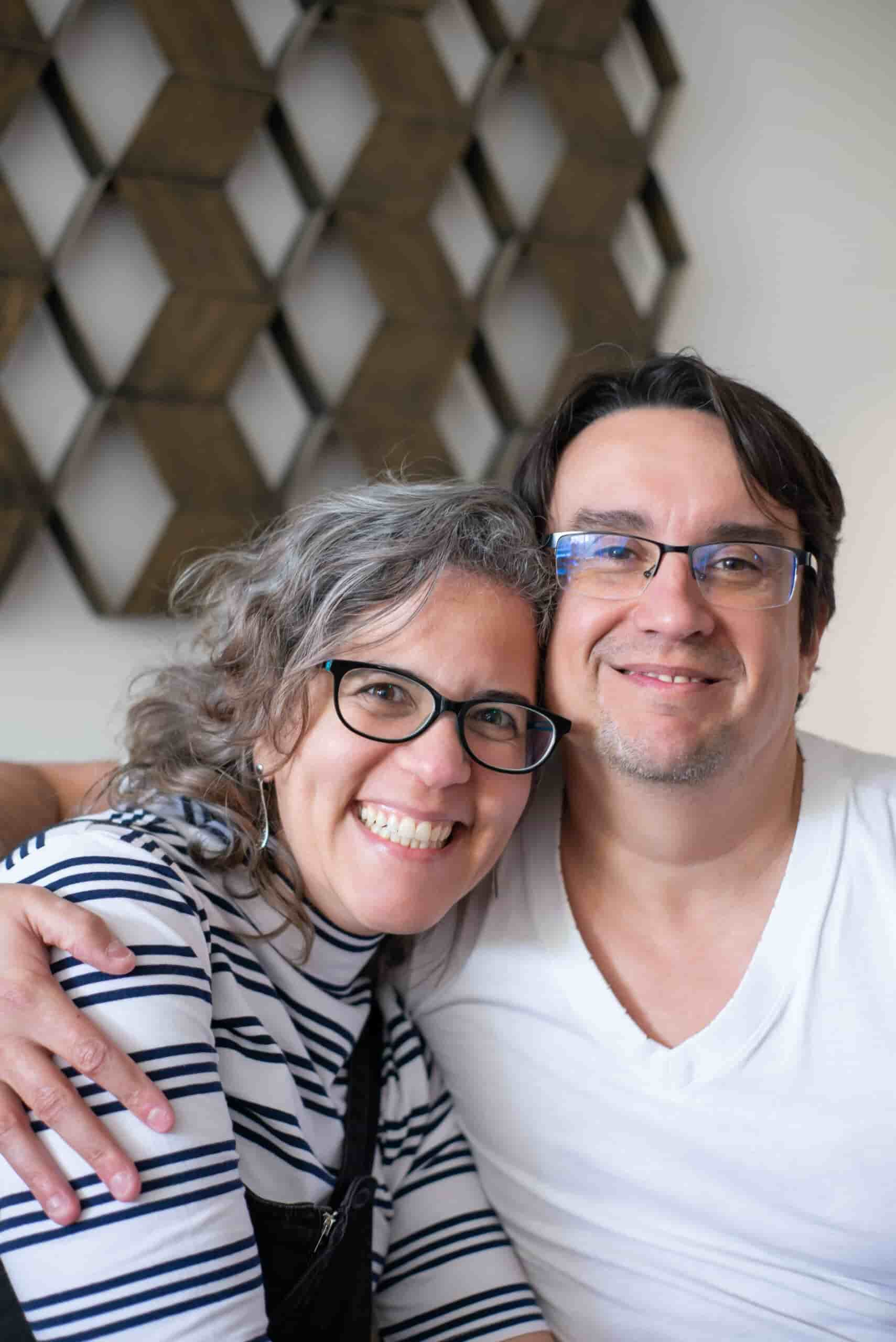 A smiling couple sits closely, with the woman wearing glasses and a striped shirt, and the man in a white shirt and glasses. They are in a cozy setting with a geometric wall pattern in the background.