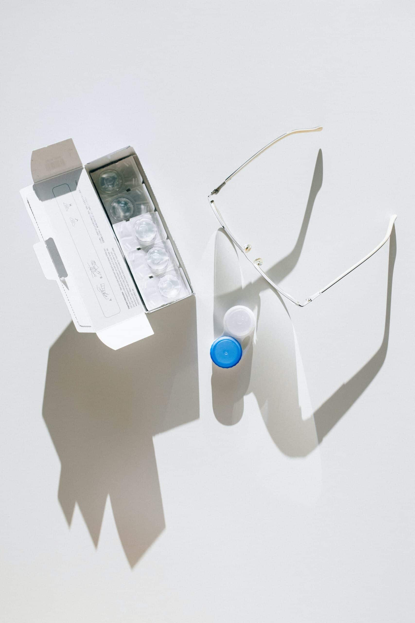 An open box of contact lenses, a lens holder with blue and white caps, and a pair of eyeglasses are placed on a light surface with shadows cast behind them.