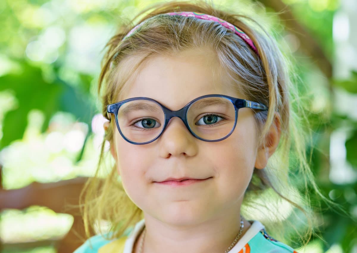 A young child with blue glasses smiles gently at the camera. The child has light brown hair held back by a headband. The background is green and blurred, suggesting an outdoor setting.