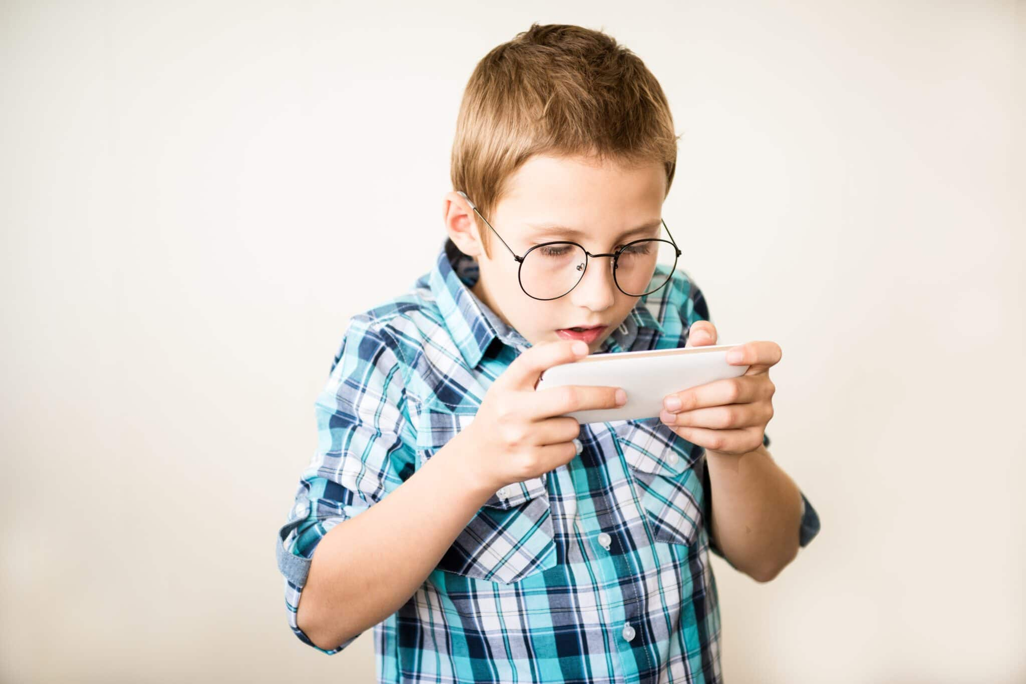 Young boy with glasses intently playing on a smartphone. He is wearing a blue plaid shirt and standing against a plain background.