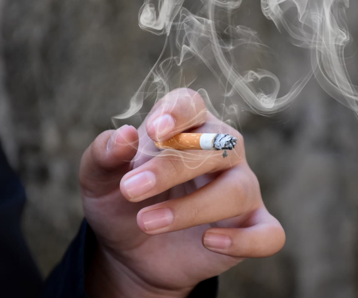 A hand holding a lit cigarette with smoke curling upwards against a blurred background.