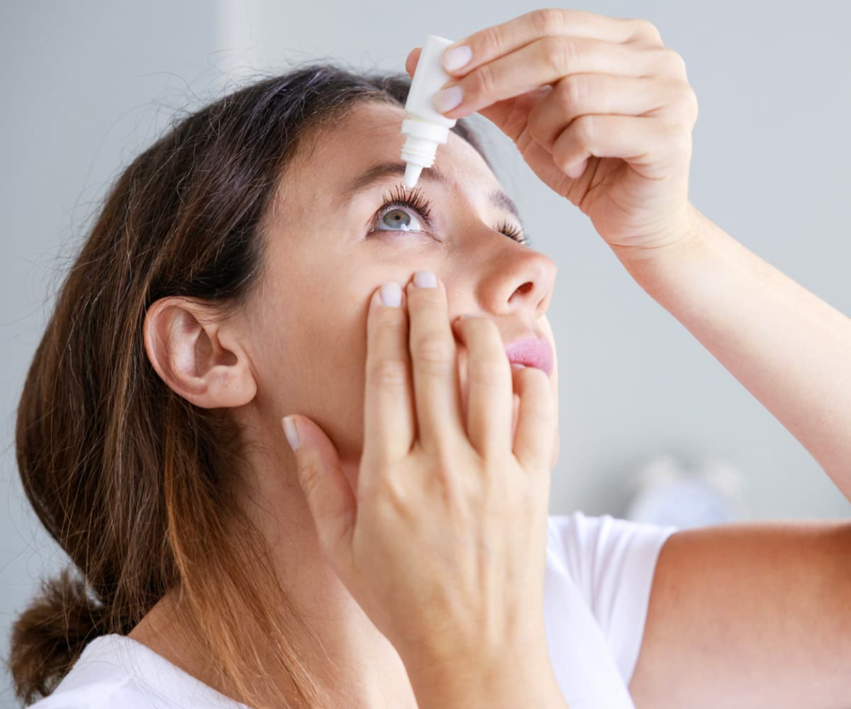 A woman with brown hair and a white shirt is tilting her head back while applying eye drops into her right eye using a small bottle. Her left hand gently pulls down her lower eyelid. She is indoors against a soft-focus background.