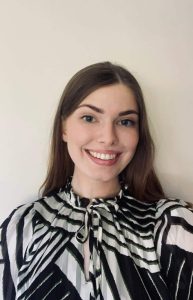 Aoife Lynch Optometric Advisor to Optometry Ireland person with long brown hair smiles at the camera. They are wearing a black and white patterned top with a bow at the neck. The background is plain white.