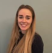 Optometrist Olivia Donegan with long blonde hair, wearing a dark top, smiles at the camera against a plain light gray background.